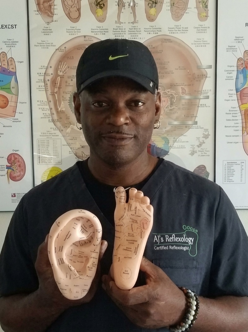 AJ Johnson, certified reflexologist, holding ear and foot reflexology models in front of a reflexology wall chart